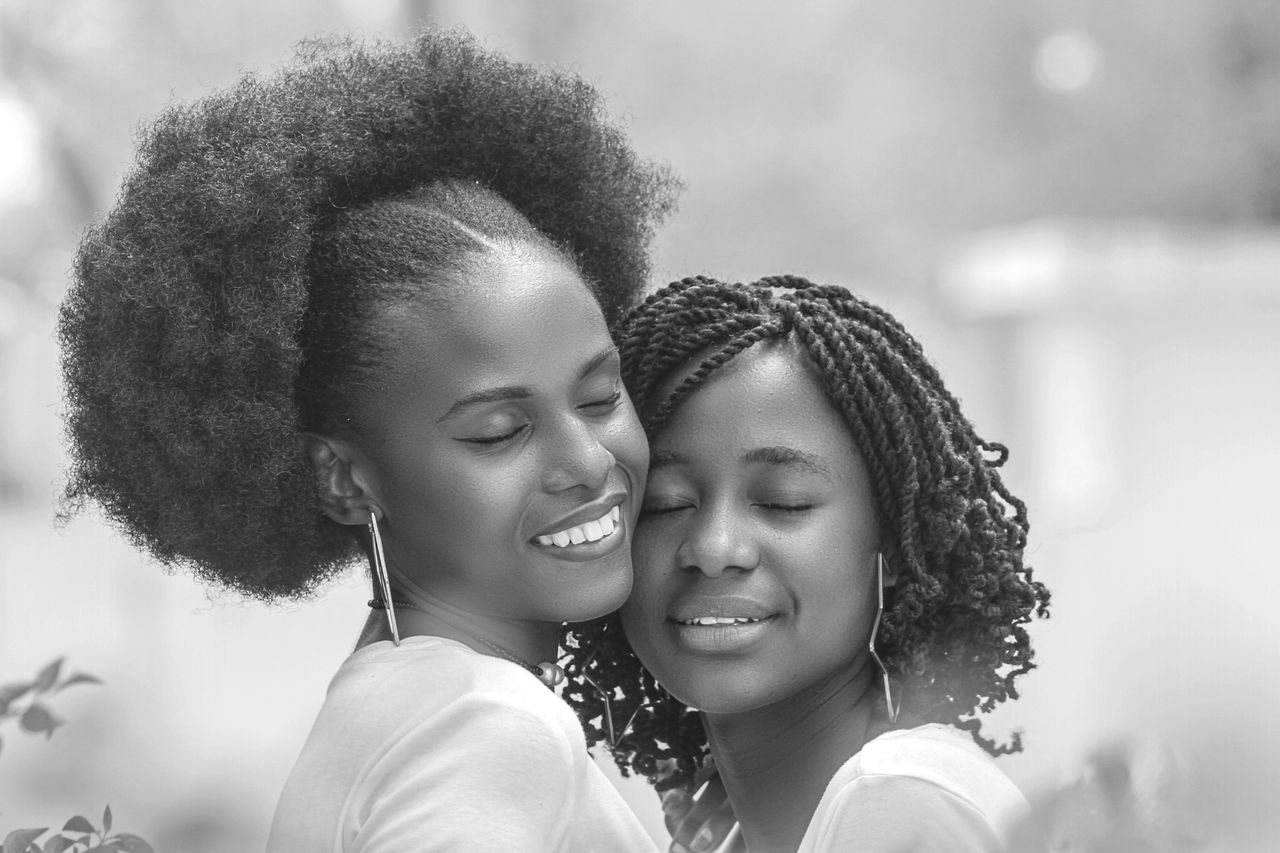Black and white of cheerful African American homosexual girlfriends with creative hairstyle embracing with closed eyes
